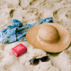 Red JBL GO3 Bluetooth speaker lying in the sand next to a towel, hat and pair of shades